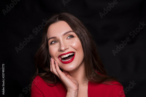 Emotional head shot portrait of a brunette caucasian woman in red dress and with red lips on black background. She smiling with hands near her face