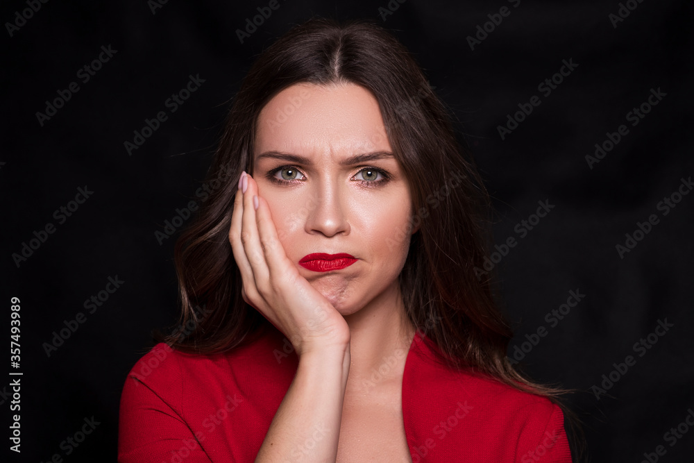 Emotional head shot portrait of a brunette caucasian woman in red dress and with red lips on black background. She have a toothache with hand on her cheek Emotional head shot portrait of a brunette caucasian woman in red dress and with red lips on black background. She have a toothache with hand on her cheek