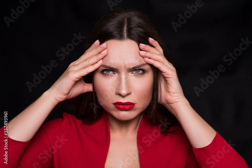 Emotional head shot portrait of a brunette caucasian woman in red dress and with red lips on black background. She have a headache, with fingers on her head