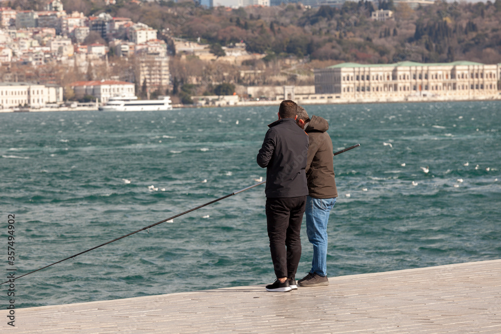 Unrecognizable two fishermen try to catch fish by Bosphorus. It is a sunny winter day in Istanbul. European side and dolmabahce palace are visible in the background. 