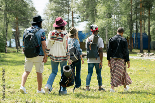 Rear view of young multi-ethnic friends with satchels walking over festival campsite while finding place for camping