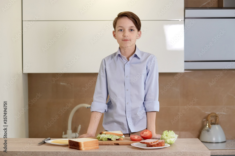 A boy stands in the kitchen in front of the table and prepares a sandwich.
