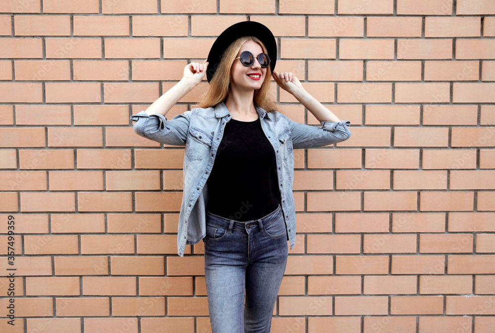 Stylish young woman wearing a black round hat, jeans jacket, female model looking away on brick wall background