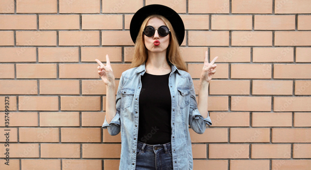 Stylish young woman blowing red lips sending sweet air kiss wearing a black round hat, jeans jacket, female model on brick wall background Stylish young woman blowing red lips sending sweet air kiss wearing a black round hat, jeans jacket, female model on brick wall background