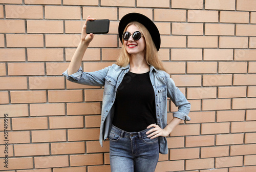 Happy smiling young woman taking selfie picture by smartphone wearing a black round hat, jeans jacket on brick wall background