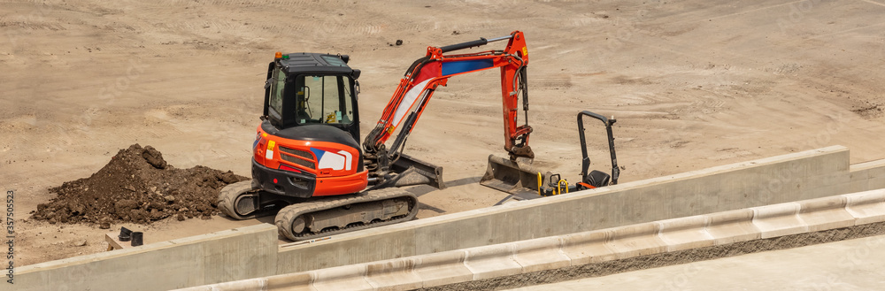 Orange excavator parked next to a concrete platform. Construction site. Aerial angled view. Orange excavator parked next to a concrete platform. Construction site. Aerial angled view.