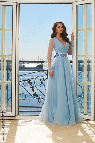 Young beautiful stunning girl in long blue dress posing in interior with open door and view on sunny blue sky. Woman in elegant ball gown