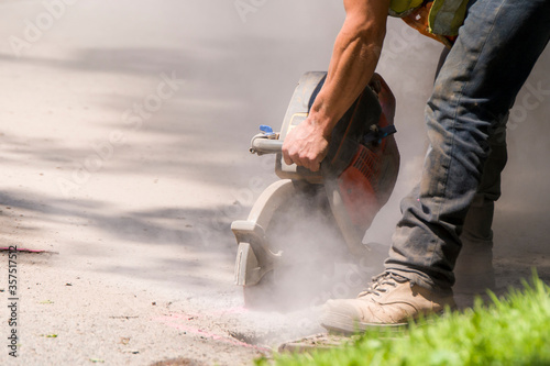 Asphalt cutter. A worker cuts through an asphalt road with a large handheld rotary cutter. Lots of dust from cutting. Worker not identifiable, only arm and leg visible. Room for text.