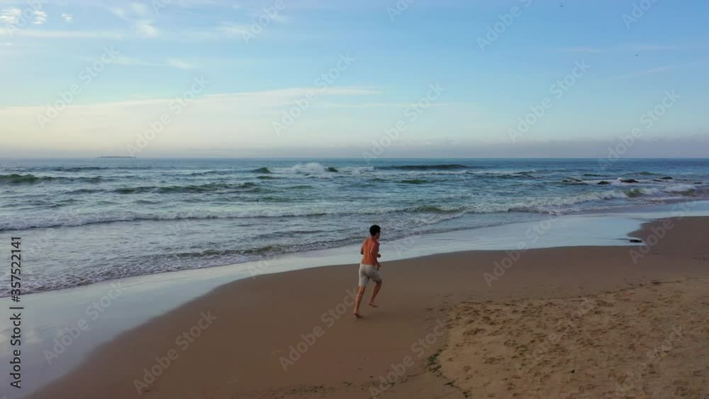 Aerial: Drone following shirtless man jogging on shore at beach against sky during sunset - Punta del Este, Uruguay Aerial: Drone following shirtless man jogging on shore at beach against sky during sunset - Punta del Este, Uruguay