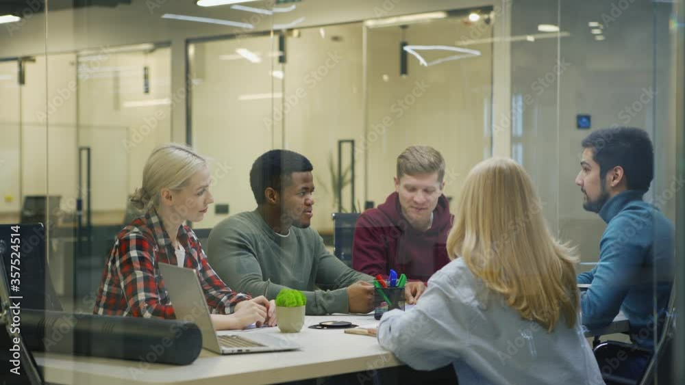 Tracking medium shot of diverse business team of five creative people brainstorming in meeting sitting at table behind glass wall in office Tracking medium shot of diverse business team of five creative people brainstorming in meeting sitting at table behind glass wall in office