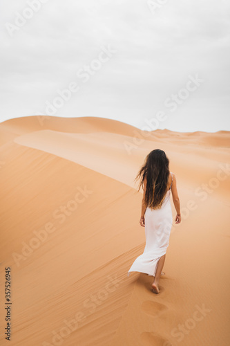 Woman with long hair in silk white dress walking in Sahara desert sand dunes in Morocco. View from the back.