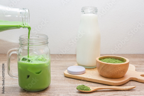 Pouring iced matcha green tea latte​ from​ buttle into mug with green tea powder and fresh milk in bottle on wooden table