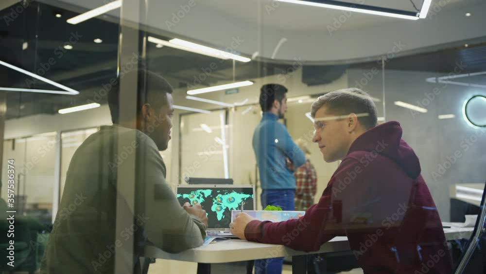 Tracking medium shot of Black young man and his Caucasian colleague sitting at desk with open laptop with world map on screen and discussing global marketing strategy behind glass wall in office Tracking medium shot of Black young man and his Caucasian colleague sitting at desk with open laptop with world map on screen and discussing global marketing strategy behind glass wall in office