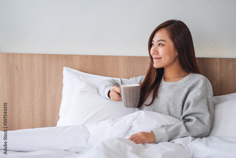 A beautiful asian woman drinking hot coffee while lying on a white cozy bed at home in the morning A beautiful asian woman drinking hot coffee while lying on a white cozy bed at home in the morning
