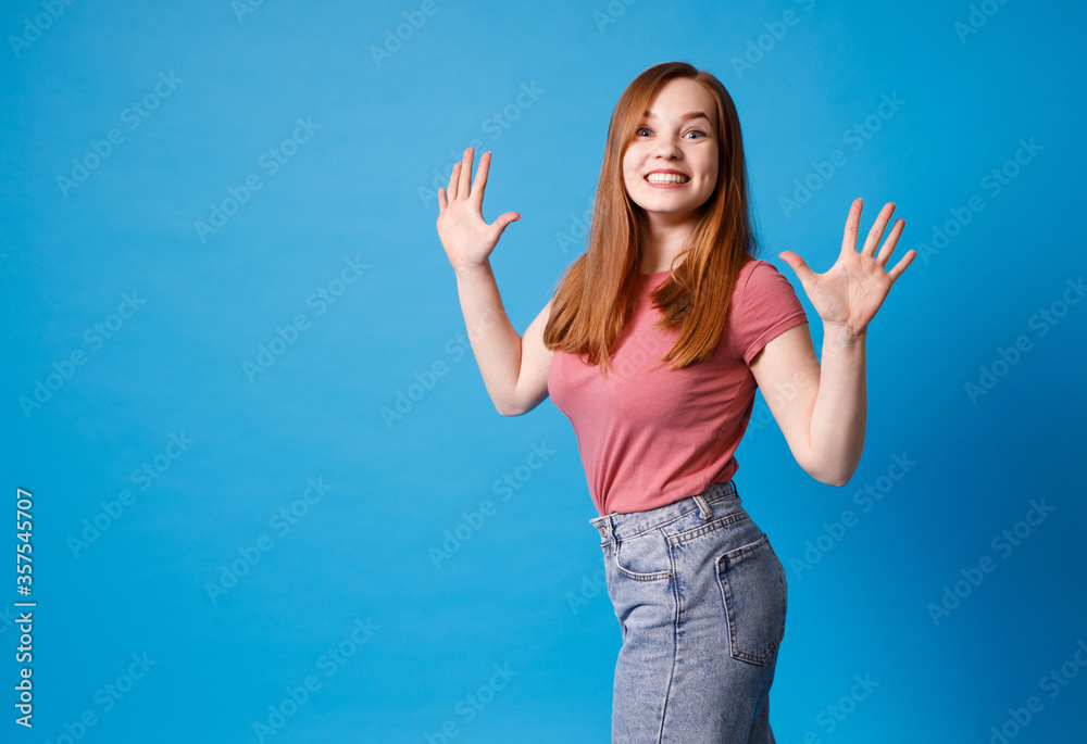 Young ginger girl in pink t-shirt and jeans posing isolated on blue background. Girl raised her hands, admiring gesture. Young ginger girl in pink t-shirt and jeans posing isolated on blue background. Girl raised her hands, admiring gesture.