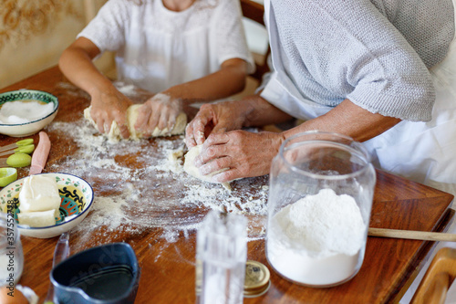 Cropped image of the hands of an old woman and a child kneading dough, top view of the dough. Ingredients and utensils for baking arranged on a wooden table.
