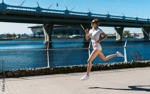 Young fitness woman runner running in the morning on the promenade or embankment and doing sports exercises. Healthy lifestyle, street training, social distancing