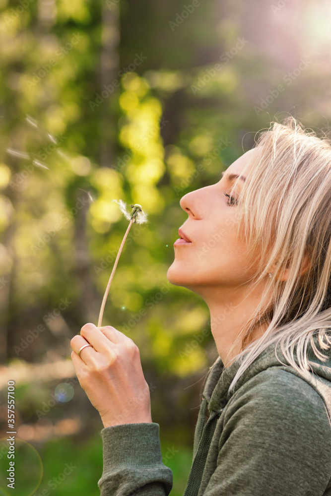 Blonde woman blowing dandelion blossom fluffs in the background of blurred dense green forest during summer evening time and illuminated by sun beams and lens flare Blonde woman blowing dandelion blossom fluffs in the background of blurred dense green forest during summer evening time and illuminated by sun beams and lens flare
