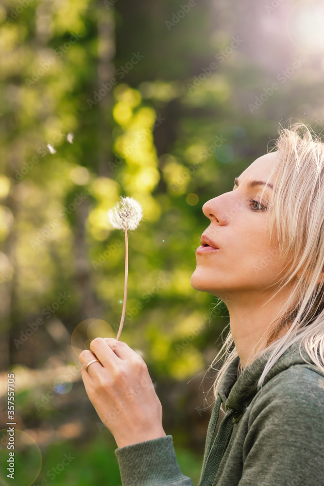 Blonde woman blowing dandelion blossom fluffs in the background of blurred dense green forest during summer evening time and illuminated by sun beams and lens flare Blonde woman blowing dandelion blossom fluffs in the background of blurred dense green forest during summer evening time and illuminated by sun beams and lens flare