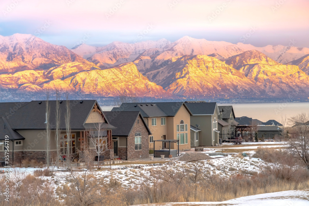Facade of homes with snowy sunlit mountain and calm lake background at sunset