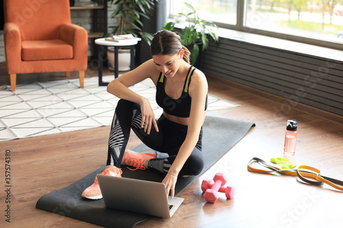 Fototapeta Naklejka Na Ścianę i Meble -  Beautiful slim sporty woman in sportswear is sitting on the floor with dumbbells and bottle of water and is using a laptop at home in the living room. Healthy lifestyle. Stay at home activities.
