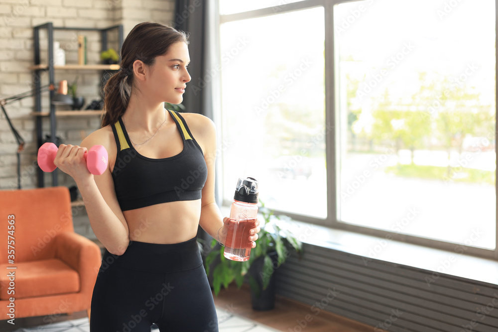 Young beautiful woman standing with dumbbell and drink water after exercising. Attractive female bodybuilder working out. Fitness and healthy lifestyle concept.