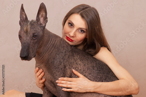 Young attractive woman embracing Mexican Hairless dog, or Xoloitzcuintle breed. Female with dark hair, eyes and red lipstick, smiling, beautiful pet with attentive look and irokez on head. photo