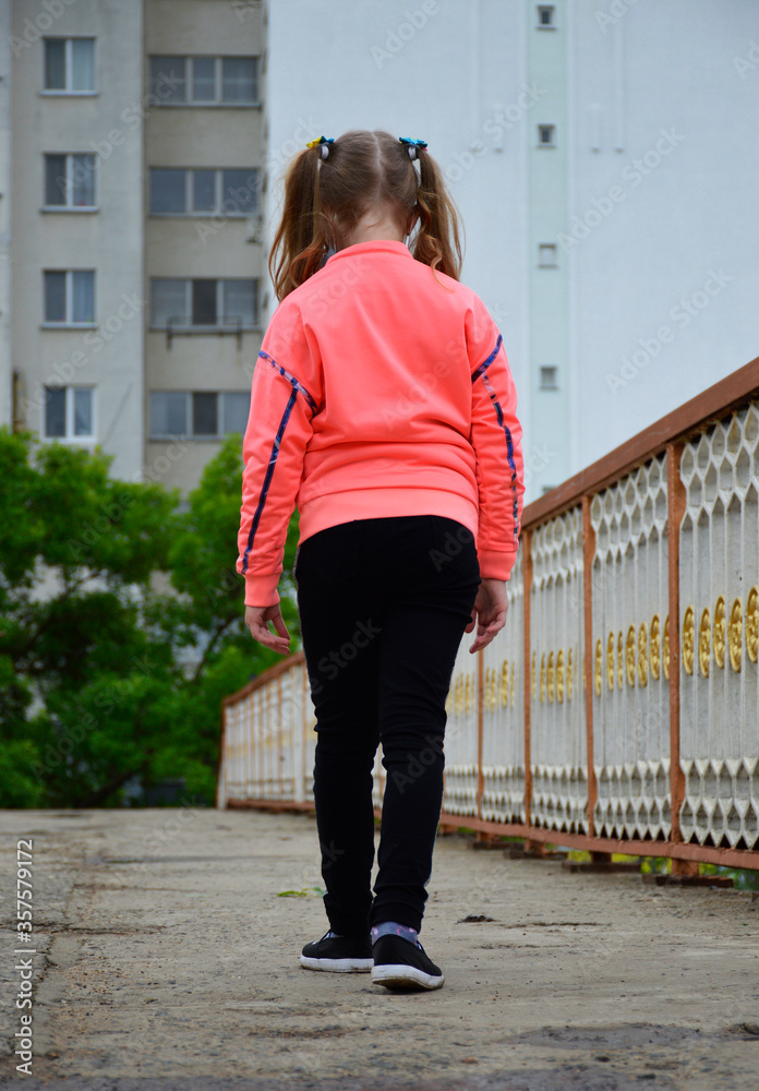 girl goes along the path of the bridge. railing on the right, in the background multi-storey buildings