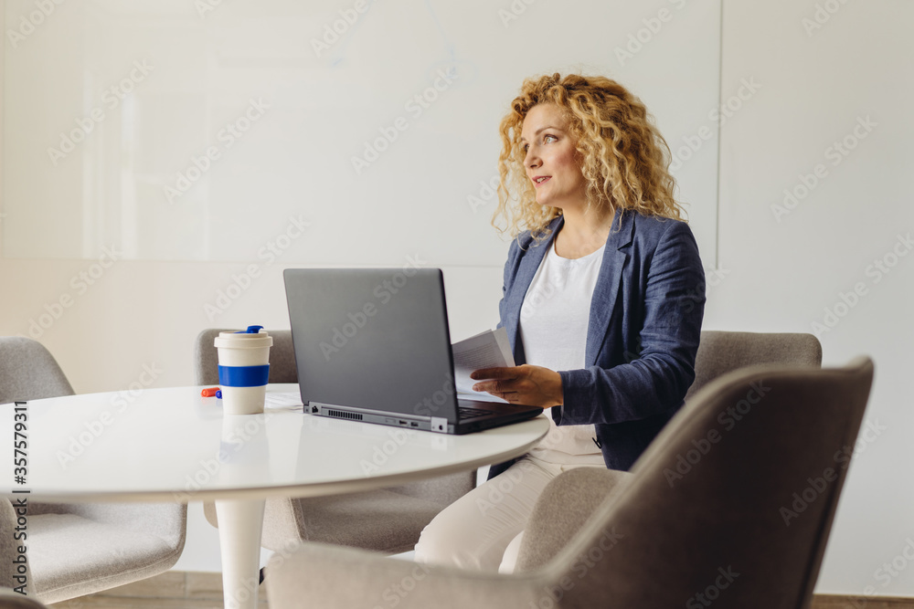 Shot of an attractive middle age blond curly business woman working on laptop in her workstation or meeting room in cozy office interior. Shot of an attractive middle age blond curly business woman working on laptop in her workstation or meeting room in cozy office interior.