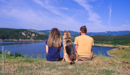 A German shepherd sits next to a guy and a girl and looks at the lake