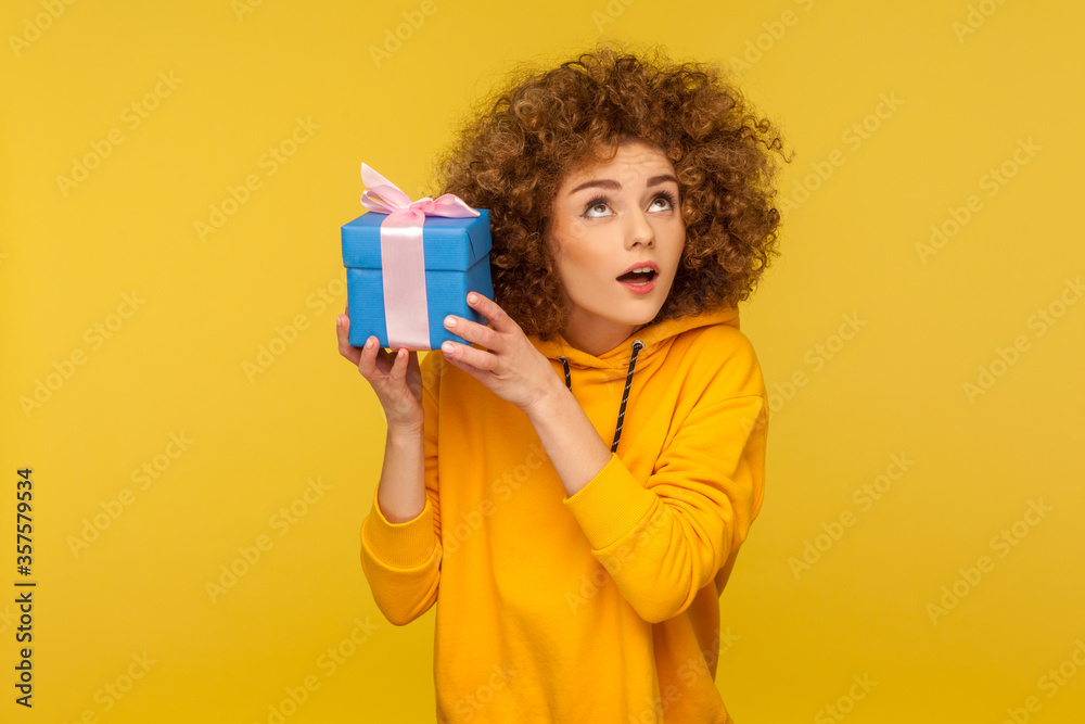 Portrait of funny curious curly-haired woman in urban style hoodie holding present box near ear and listening what's inside, in anticipation of dream gift. studio shot isolated on yellow background