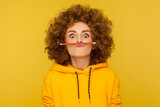 Closeup of comical curly-haired young woman in urban style hoodie holding pencil with her lips, pretending to have mustache and looking with amazed eyes. studio shot isolated on yellow background