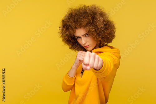 Struggle and fighting spirit. Portrait of aggressive curly-haired woman in urban style hoodie attacking with clenched fists, threatening to punch. indoor studio shot isolated on yellow background