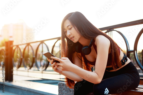 Asian girl is listening to music on headphones. Girl on a walk. Summer time. In the hands of the phone. Young girl during sunset. Girl in a sports bow sits on a bench.