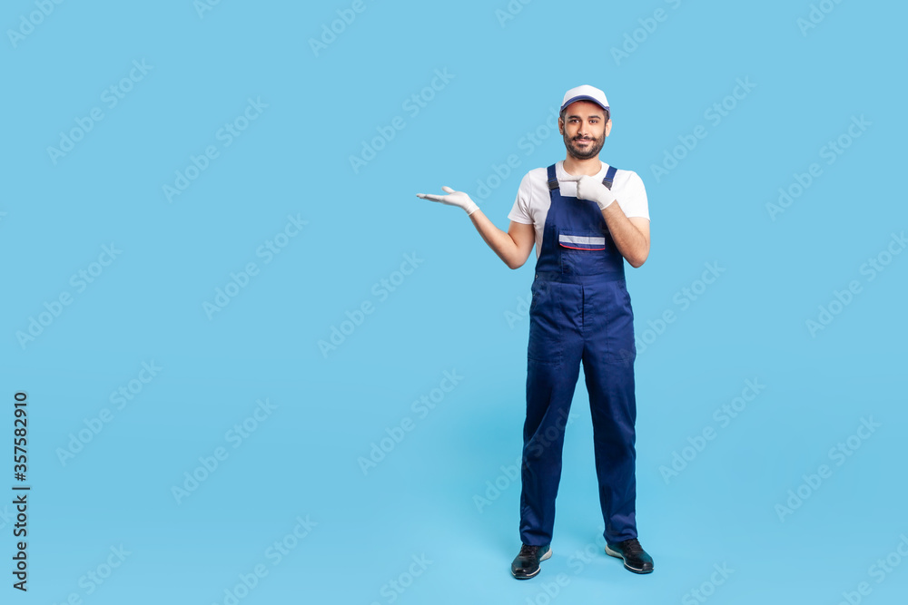 Naklejka premium Full length cheerful worker in blue overalls, cap and protective gloves standing, pointing at empty space on his hand. Profession of service industry, courier or labourer. studio shot isolated