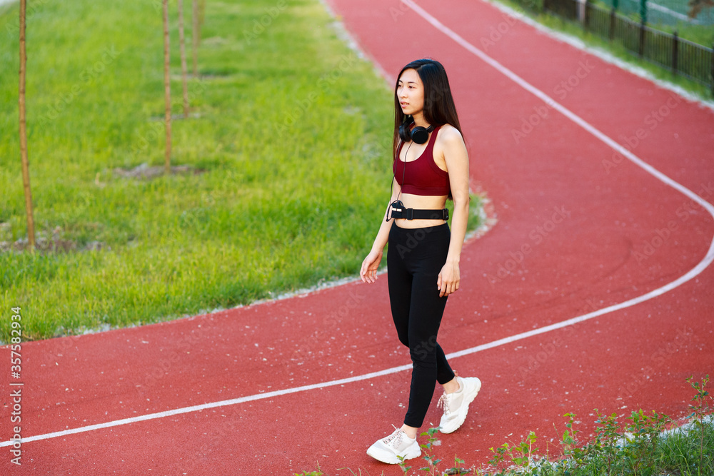 Asian girl go in for sports on a sports track on the street. Red treadmill. Girl with headphones. Girl in a sports top and leggings.