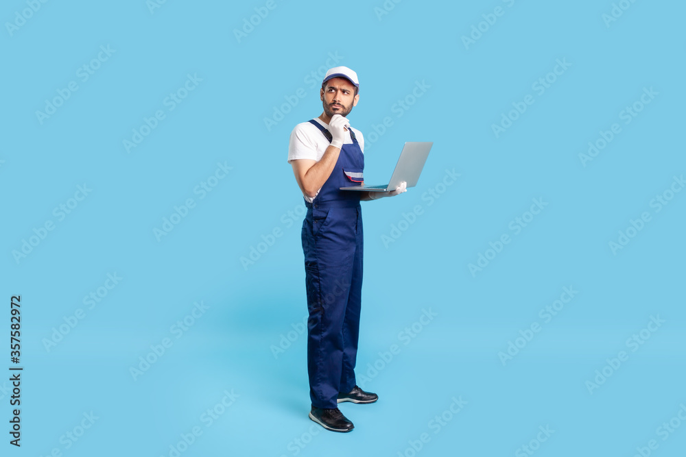Full length thoughtful uncertain workman in uniform and protective gloves holding laptop, pondering decision with doubtful expression, online order for repair services. indoor studio shot isolated