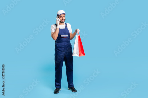 Full length portrait of smiling courier in workwear talking on cellphone, holding shopping bags and communicating with client. Delivery man answering phone call, accepting order at fashion store