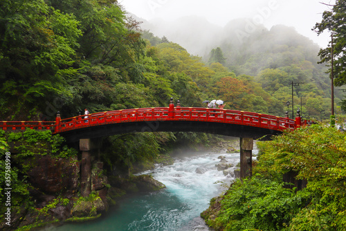People with umbrellas over red bridge Shinkyo crossing Daiya river. Rainy day, magical and mysterious atmosphere. Beginning of autumn, moth of October. Nikko, Japan, Asia. Horizontal picture