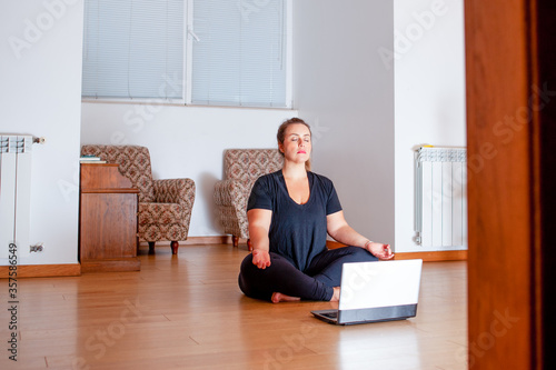 Plus size woman practicing yoga in her living room. She is watching video lessons on her computer. Copy space