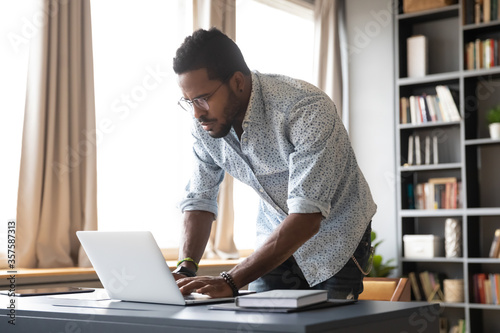 Serious African man standing in modern office room or home leaned over laptop typing answer to client, browse internet website, hurry up finish task using computer. Workday modern tech usage concept