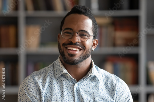 Head shot of african american bearded guy with pierced ear casual shirt smiling looking at camera standing indoor. Webcam view, conference video call, confident company representative portrait concept