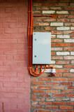 Metal electrical panel on a brick wall. It is connected by wires in orange corrugation. The wall is half painted pink, the second part is made of red brick.