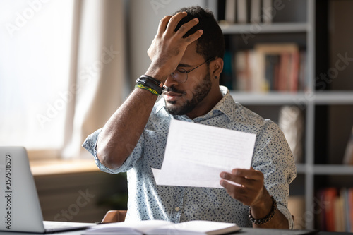 Stressed african businessman sit at desk hold paper letter reading bad awful negative news got fired from work. Dismissal notice, financial problems debt notification from bank, unpaid invoice concept photo