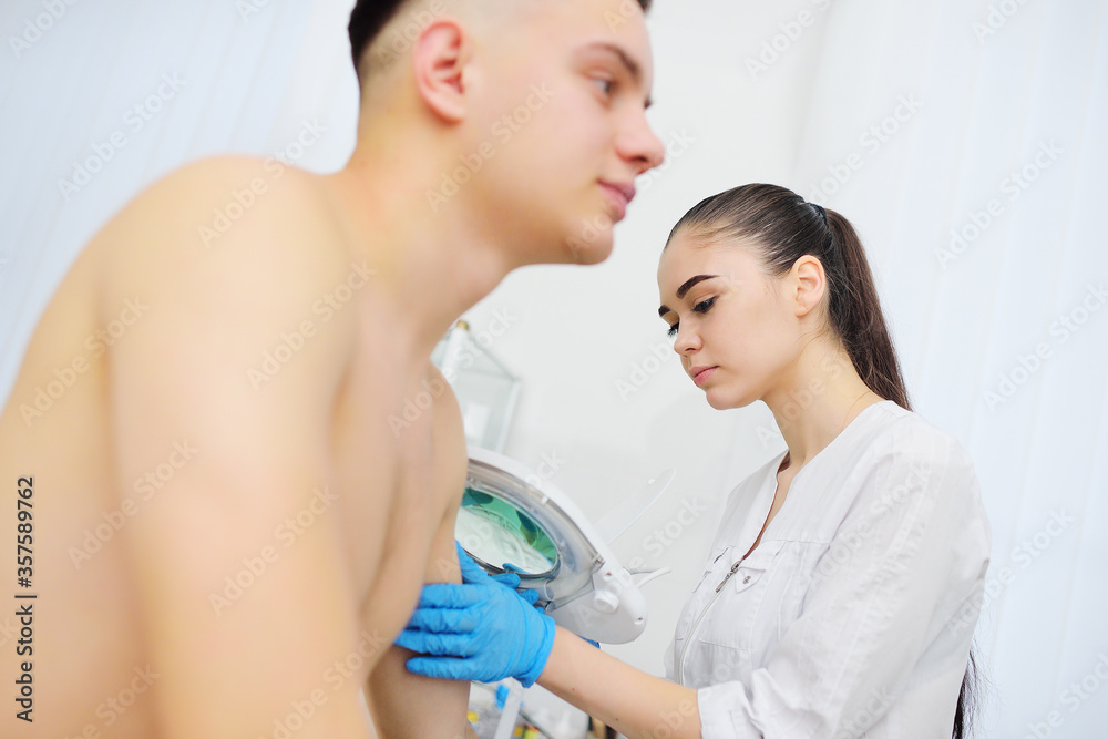 Fototapeta premium a young woman doctor dermatologist cosmetologist examines the skin structure of a male patient through a cosmetic magnifier lamp. Prevention of melanoma and skin cancer. Moles, scars, 