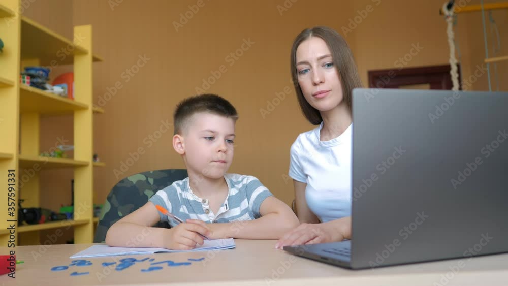 Young mother sitting at a table at home helping her son with his homework from school Young mother sitting at a table at home helping her son with his homework from school