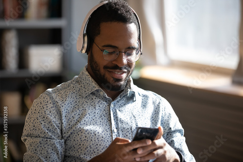 African man wear headphones holds cellphone choose track listens digital music or podcast, using video streaming service. Language learning platform most downloaded education app advertisement concept