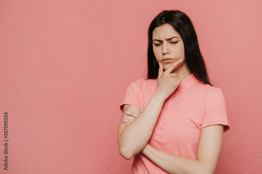 Pensive displeased woman with, holds chin and looks away, contemplates about something, wears pink t shirt, being in upset mood, smirks face and being upset by bad thoughts Pensive displeased woman with, holds chin and looks away, contemplates about something, wears pink t shirt, being in upset mood, smirks face and being upset by bad thoughts