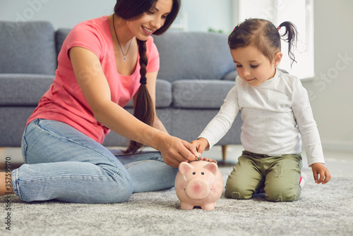 Fotografie Young mother and her daughter putting coins into piggy bank at home