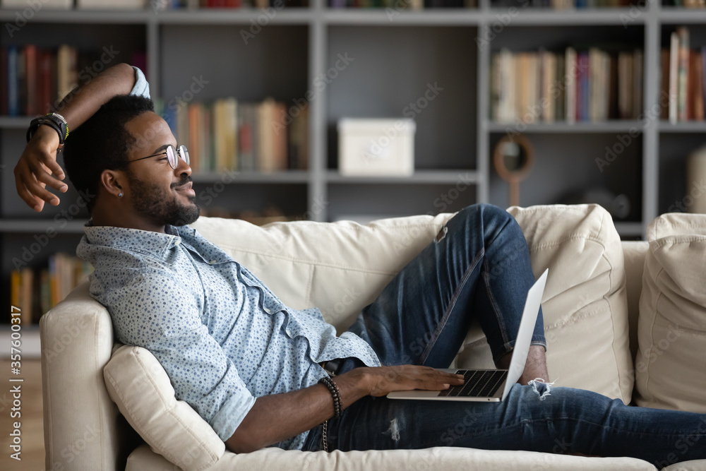 African guy relieving fatigue resting on comfy couch in modern living room put on lap notebook using media services app distracted from gadget looks at window daydreaming feels good no stress concept African guy relieving fatigue resting on comfy couch in modern living room put on lap notebook using media services app distracted from gadget looks at window daydreaming feels good no stress concept